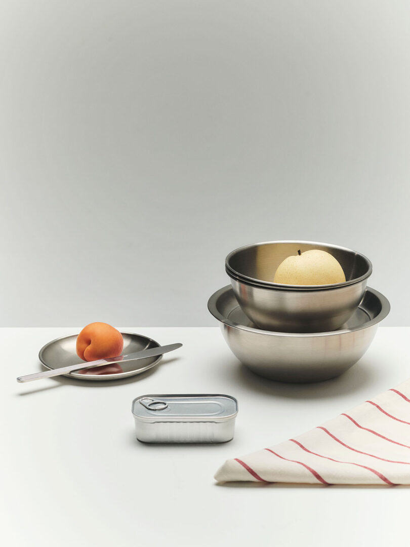A metal tin, a plate with a spoon, fork, and apricot, stacked metal bowls with a pear, and a folded white cloth with red stripes on a white surface.