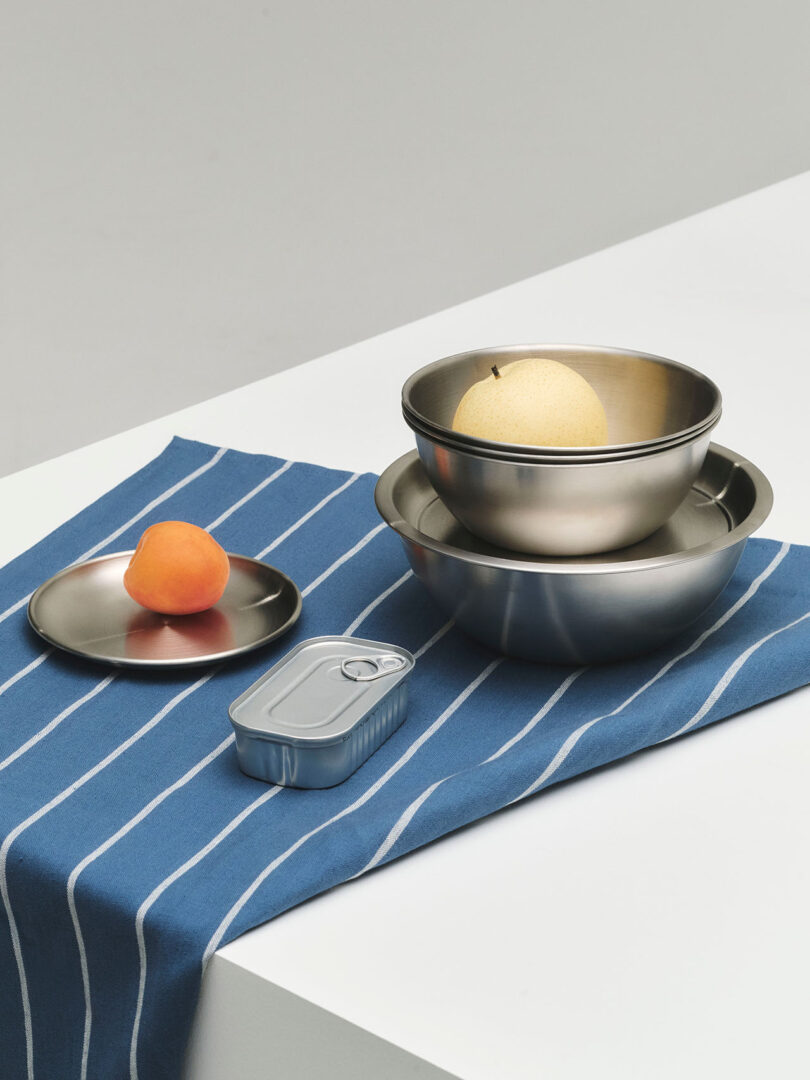 A metal plate with an apricot, a tin can, and nested stainless steel bowls with a pear on a blue striped cloth on a white table.