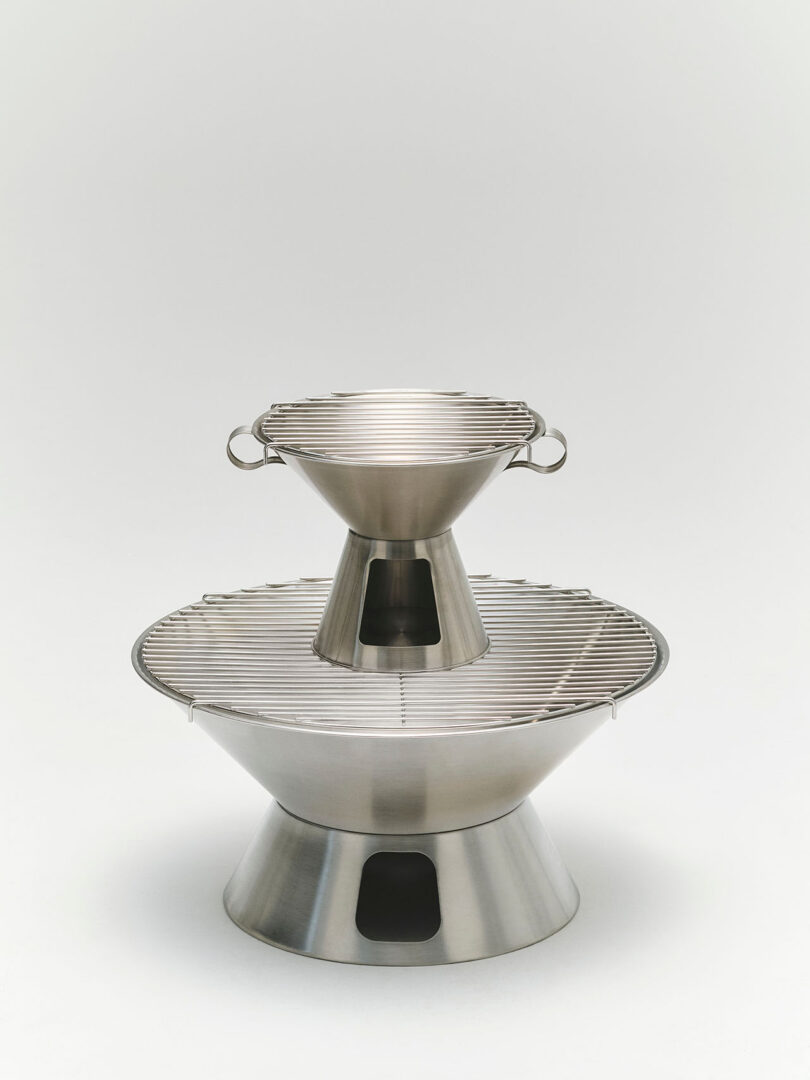 Two stainless steel grills, one small and one large, are stacked vertically. Both have a conical base and a circular grilling surface with metal handles on the smaller grill.