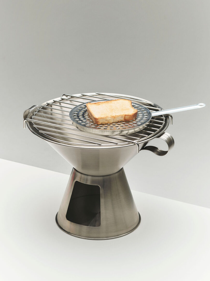 A single slice of bread sits on a spatula over a metal grill, which is placed on a stainless steel, cone-shaped barbecue grill.