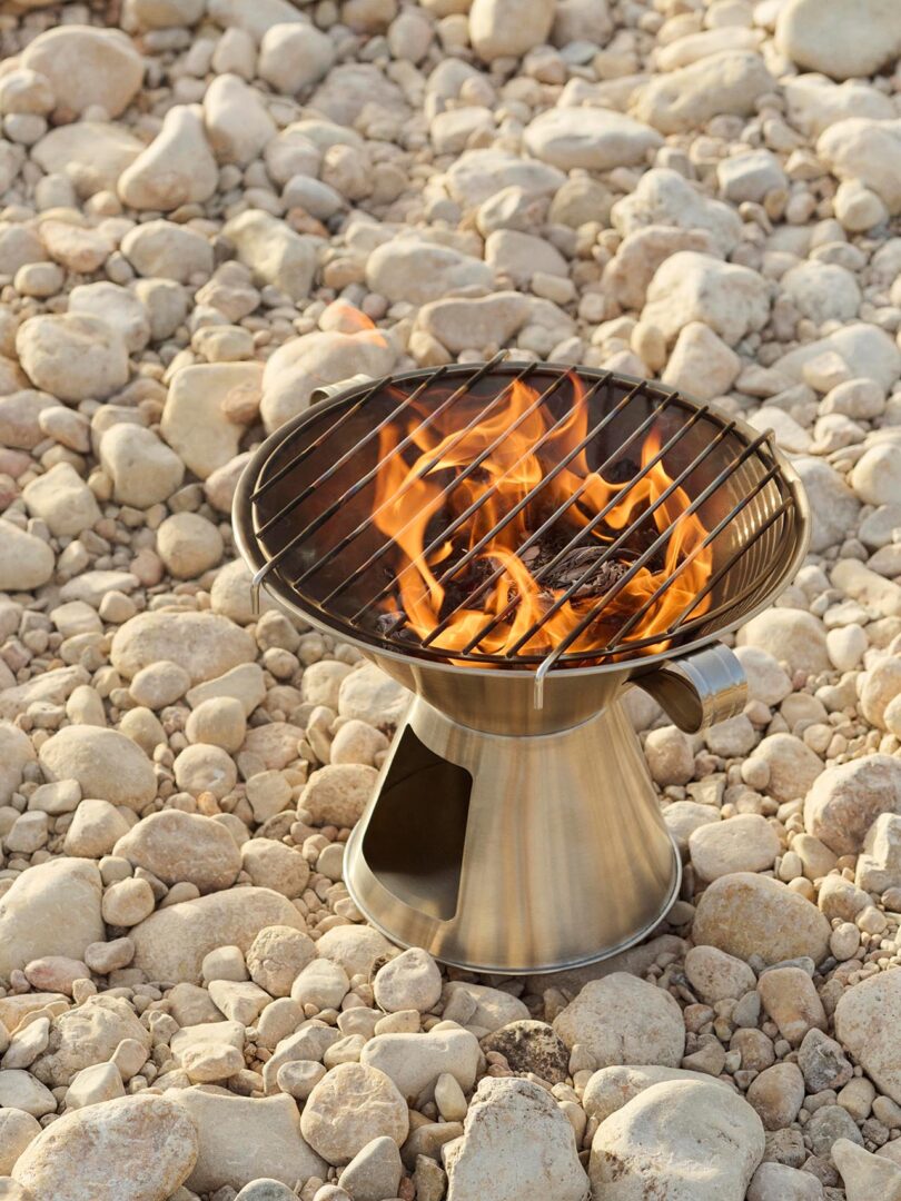 A portable metal grill with an open flame sits on a rocky surface surrounded by light-colored stones.