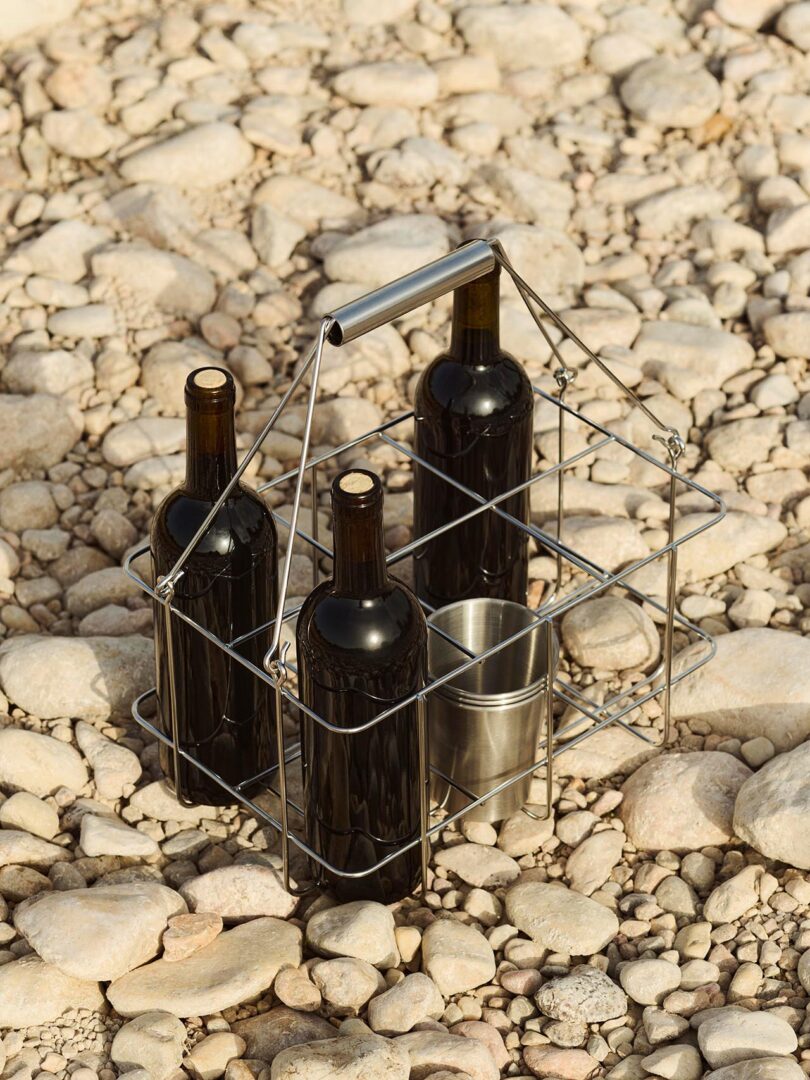 A metal carrier holding three dark wine bottles and a metal cup is placed on a rocky, pebble-covered surface.