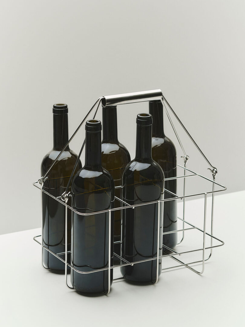 Five dark glass bottles in a metal wire carrier with a handle, placed on a white surface against a light background.