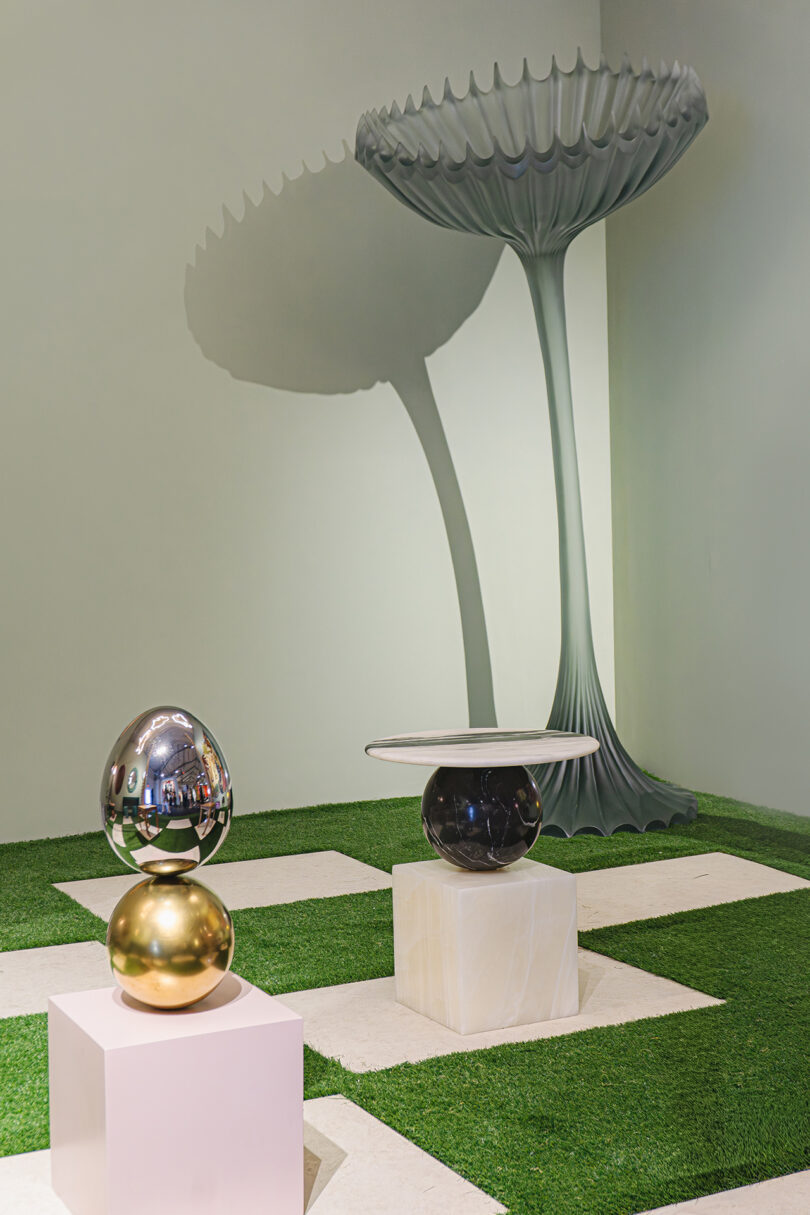 Modern art installation with reflective spheres on plinths and a large, scalloped-edged sculpture in the background, set on a checkerboard grass and stone floor.