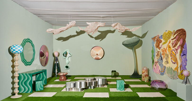 A room with pastel green walls and grass-like flooring features abstract furniture, wavy mirrors, metallic stools, hanging sculptures, and colorful artwork on the walls.