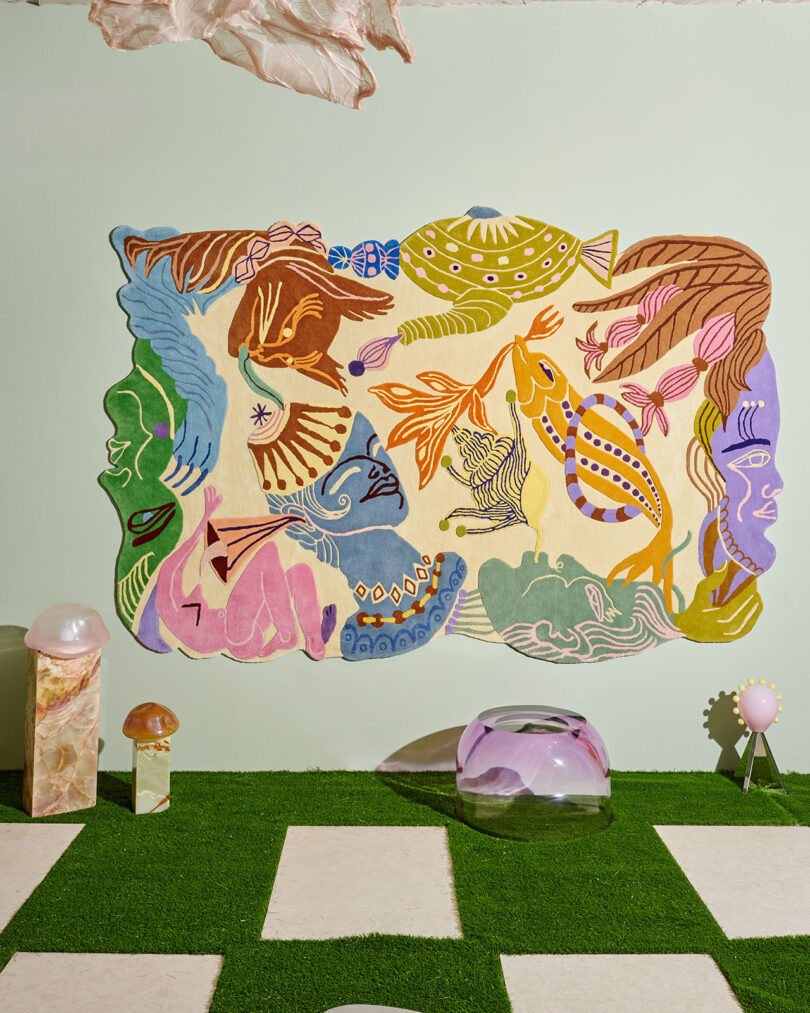 Colorful abstract wall tapestry with faces and shapes hangs on a pale wall above artificial grass, geometric tiles, and decorative objects, including mushrooms and glass domes.