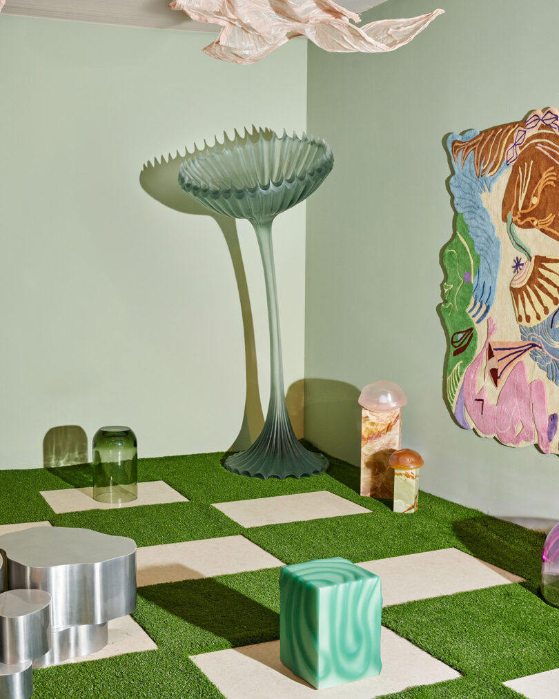 A room with artificial grass flooring, white square tiles, modern abstract furniture, a tall sculptural lamp, and a colorful wall tapestry.