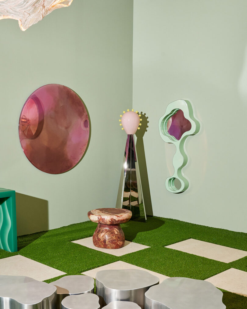 A modern room with geometric decor, including a round pink mirror, abstract green wall art, a pink and brown stool, metallic surfaces, and artificial grass flooring with white tiles.