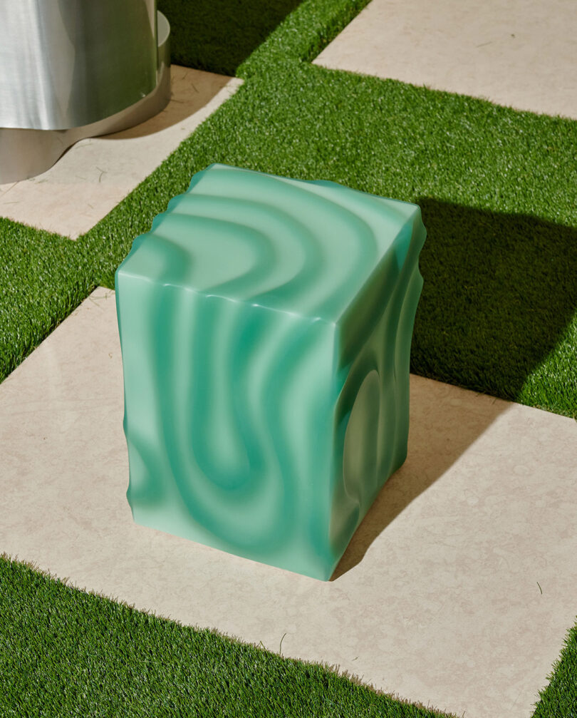 A green, wavy-patterned cube-shaped object sits on a tiled patio with artificial grass borders.