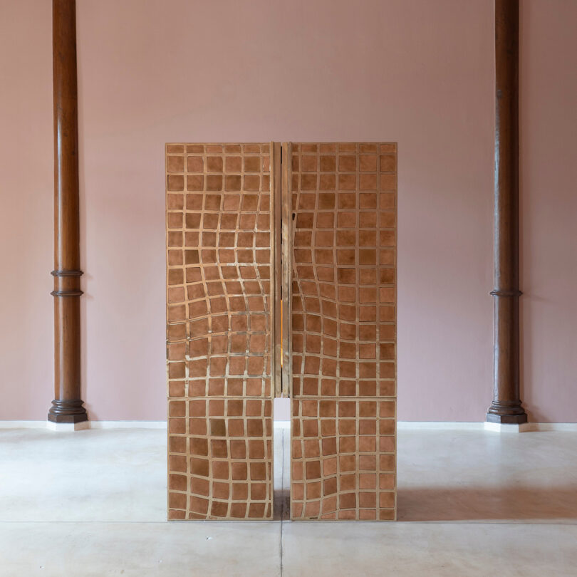 A wooden cabinet with a wavy, grid-like pattern—a nod to Kelly Wearstler’s signature style—stands in the center of a room with two wooden columns and a pink wall in the background.