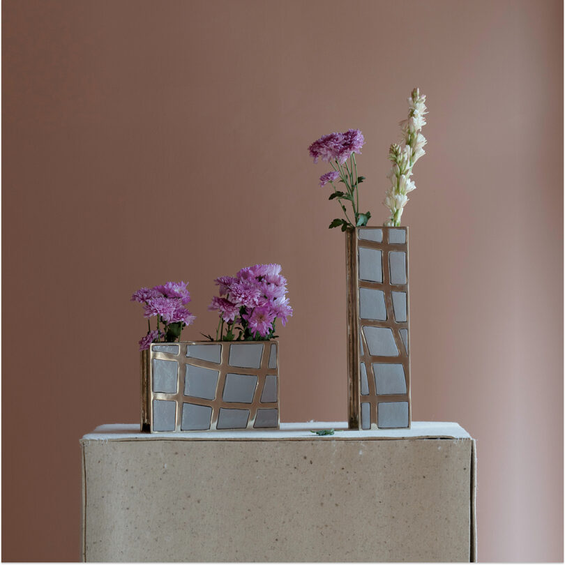 Three geometric vases with silver and gold accents, reminiscent of Kelly Wearstler's style, hold purple and white flowers, arranged on a light pedestal against a brown background.