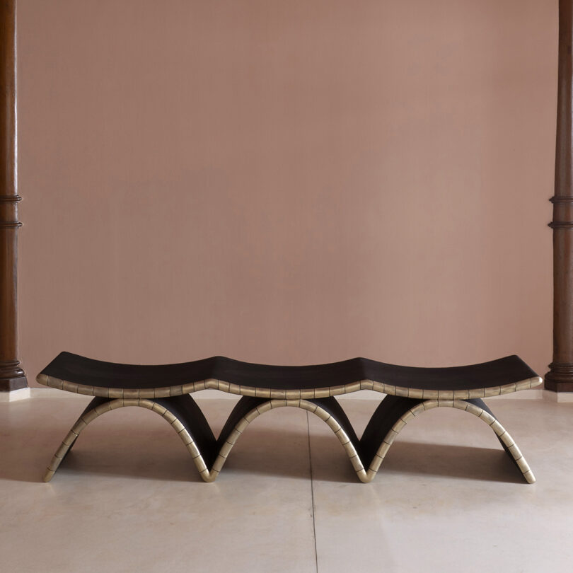 A sculptural bench with a wavy, undulating form and dark surface, supported by light-colored, curved legs, channels Kelly Wearstler’s signature style against a plain mauve wall and light floor.