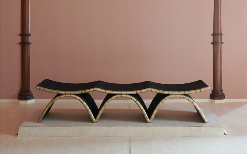 A sculptural bench with a wavy black seat and gold-edged arches, inspired by Kelly Wearstler, sits on a stone platform in a minimalist room with pink walls and two wooden columns.