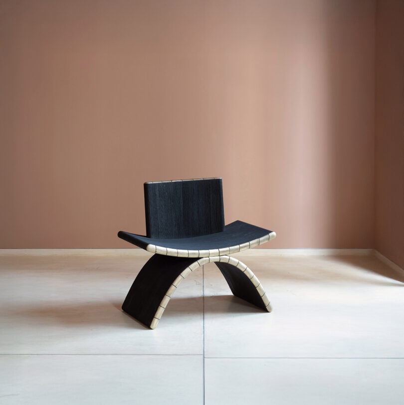 A modern black chair with a curved, arch-like base inspired by Kelly Wearstler sits on a light floor, featuring beige accents against a muted pink wall.
