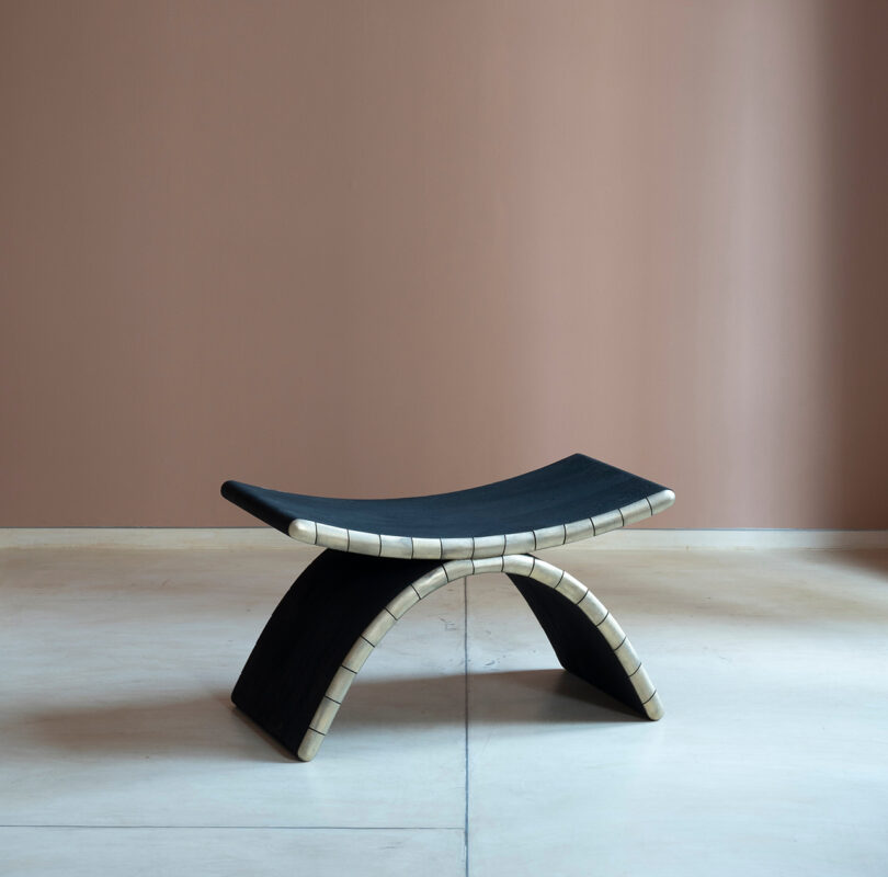 A modern black bench inspired by Kelly Wearstler, with a curved seat and arched base featuring light-colored accents, set against a pinkish-brown wall and light floor.