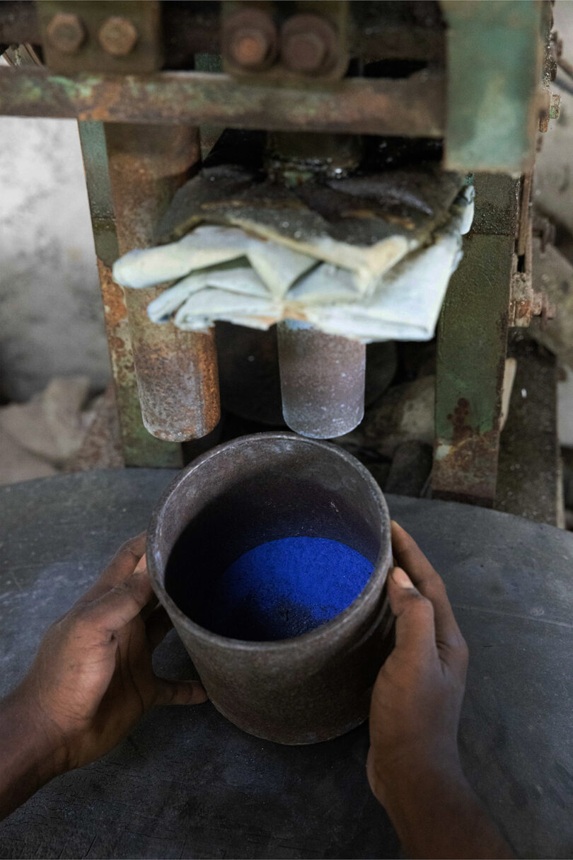 A person holds a metal container filled with blue powder under a pressing machine with folded cloths above, evoking the bold, tactile aesthetic of Kelly Wearstler.