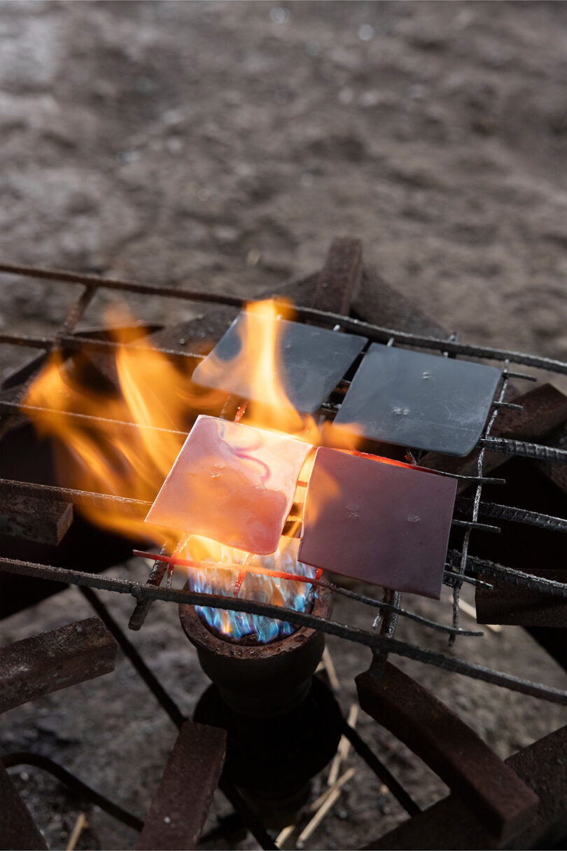 Four metal squares are heated over a gas burner with visible blue and orange-yellow flames, placed on a metal grate outdoors—evoking a bold, sculptural look reminiscent of Kelly Wearstler’s iconic design style.