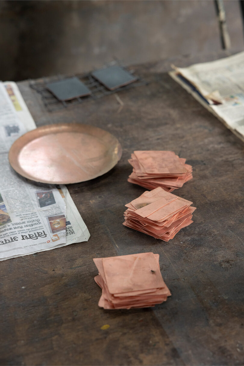 Stacks of copper sheets and a round copper plate, reminiscent of Kelly Wearstler’s bold style, are arranged on a wooden table with some newspapers nearby.