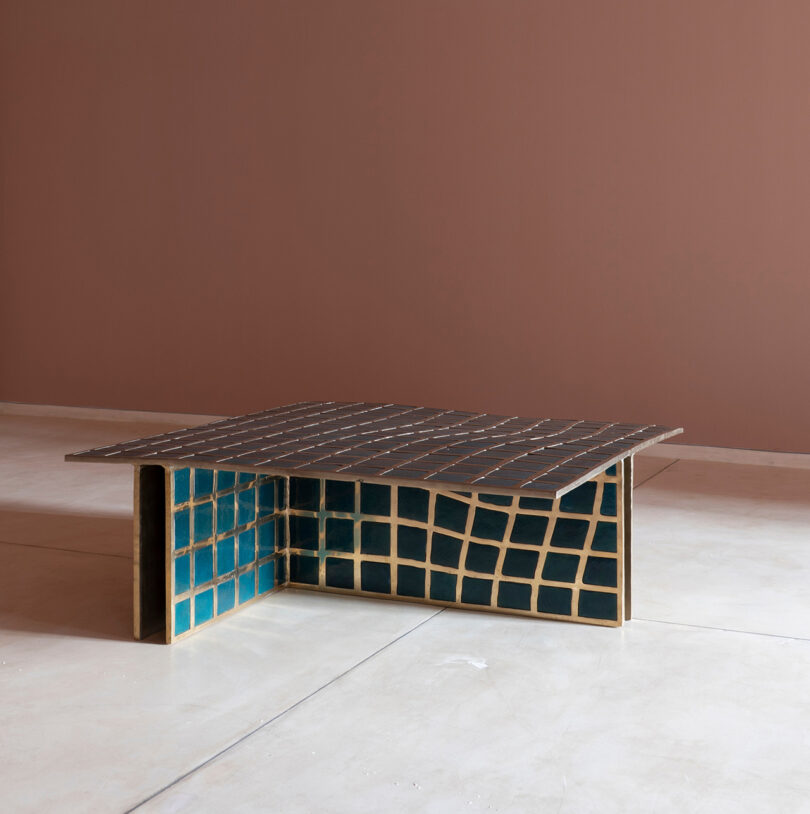 A modern coffee table with an uneven grid pattern in dark brown and gold, inspired by Kelly Wearstler, featuring blue tiled sides, set against a minimalist pinkish wall and light floor.