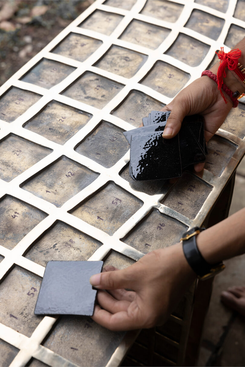 Two hands place shiny black tiles on a metal grid with numbered squares, evoking the bold, modern style of Kelly Wearstler.