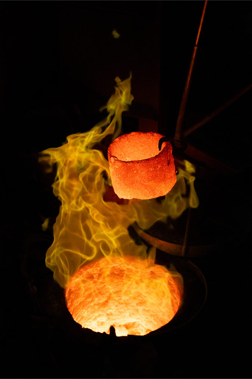 A glowing crucible filled with molten metal is lifted above a furnace, with yellow flames rising around it—a scene echoing the bold, dramatic style of Kelly Wearstler.