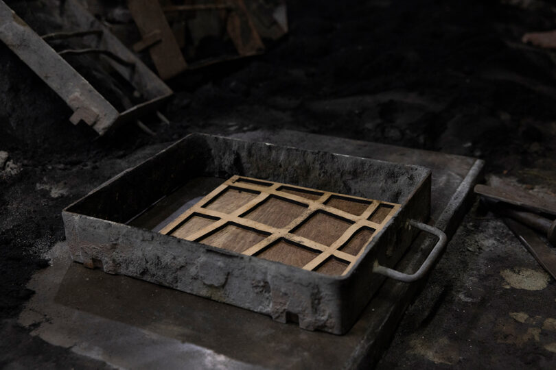 A rectangular metal tray with a wooden grid inside, reminiscent of Kelly Wearstler's bold design aesthetic, is placed on a dark, industrial surface.