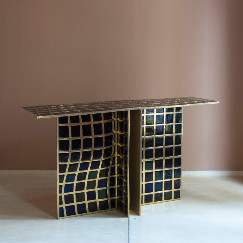 Rectangular console table with a gold metal frame and a wavy grid pattern of dark blue and gold tiles, reminiscent of Kelly Wearstler’s signature style, set against a plain beige wall and light floor.