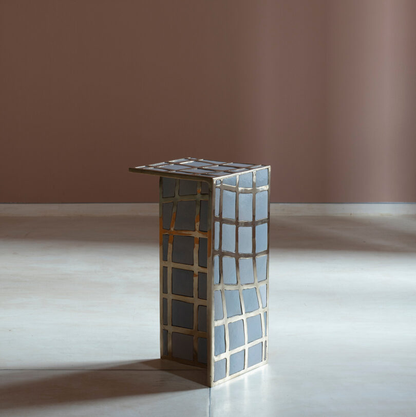 A small, modern side table inspired by Kelly Wearstler, featuring a geometric grid pattern in gray and gold, stands on a plain, light-colored floor against a muted mauve wall.