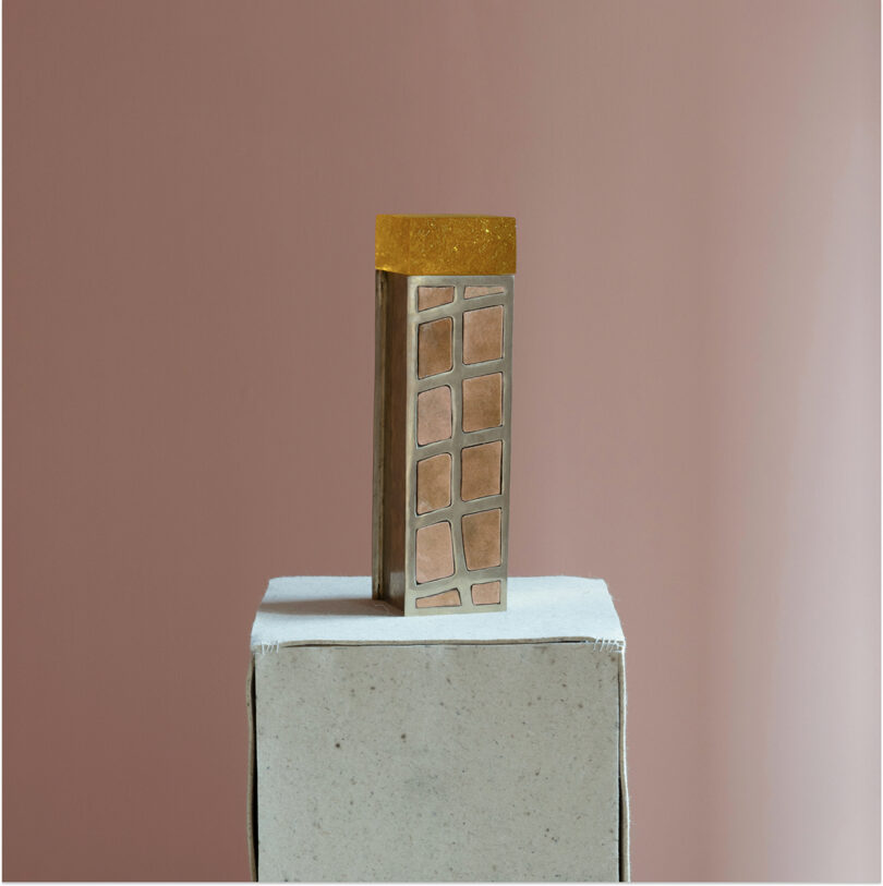 A rectangular object with a gold top and geometric metallic sides, reminiscent of Kelly Wearstler's signature style, sits on a white textured pedestal against a muted pink background.