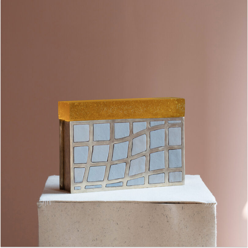 A rectangular object with a gold top and a silver, irregular grid pattern on the front, reminiscent of Kelly Wearstler's signature style, rests on a textured beige platform against a muted pink background.