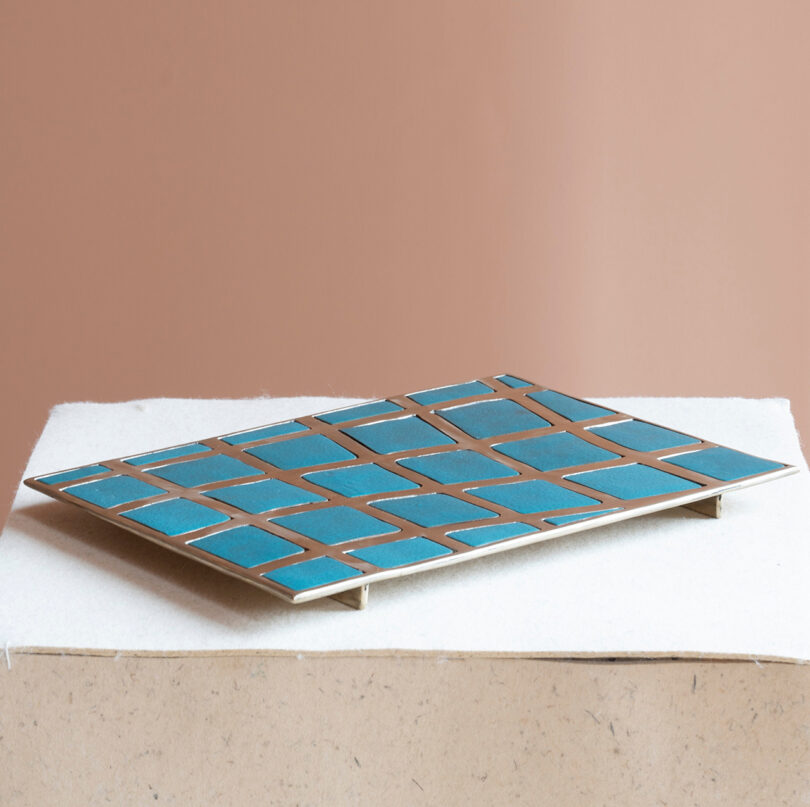A rectangular tray with a grid pattern of blue tiles and brass edges, inspired by Kelly Wearstler’s bold style, displayed on a white surface against a beige background.