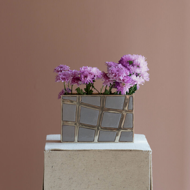 Rectangular Kelly Wearstler-inspired vase with a geometric pattern holding light and dark purple flowers, set on a fabric-covered pedestal against a plain mauve background.