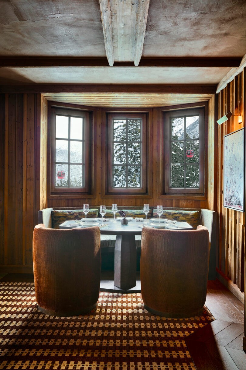 A cozy dining nook with a round table, four place settings, and two curved brown chairs in front of three large windows, with snowy trees visible outside.