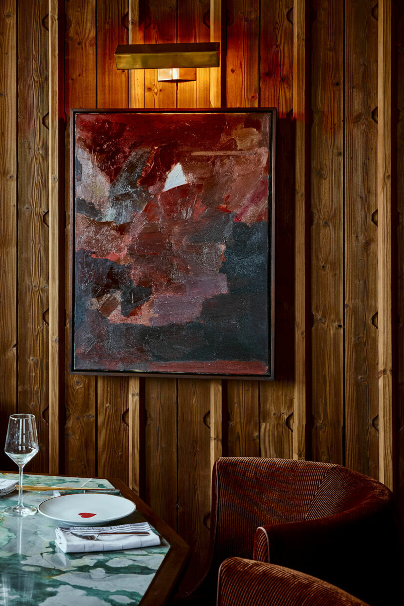A framed abstract painting with red and dark tones hangs on a wooden wall above a dining table set with a plate, wine glass, and cutlery. A brown chair is positioned beside the table.