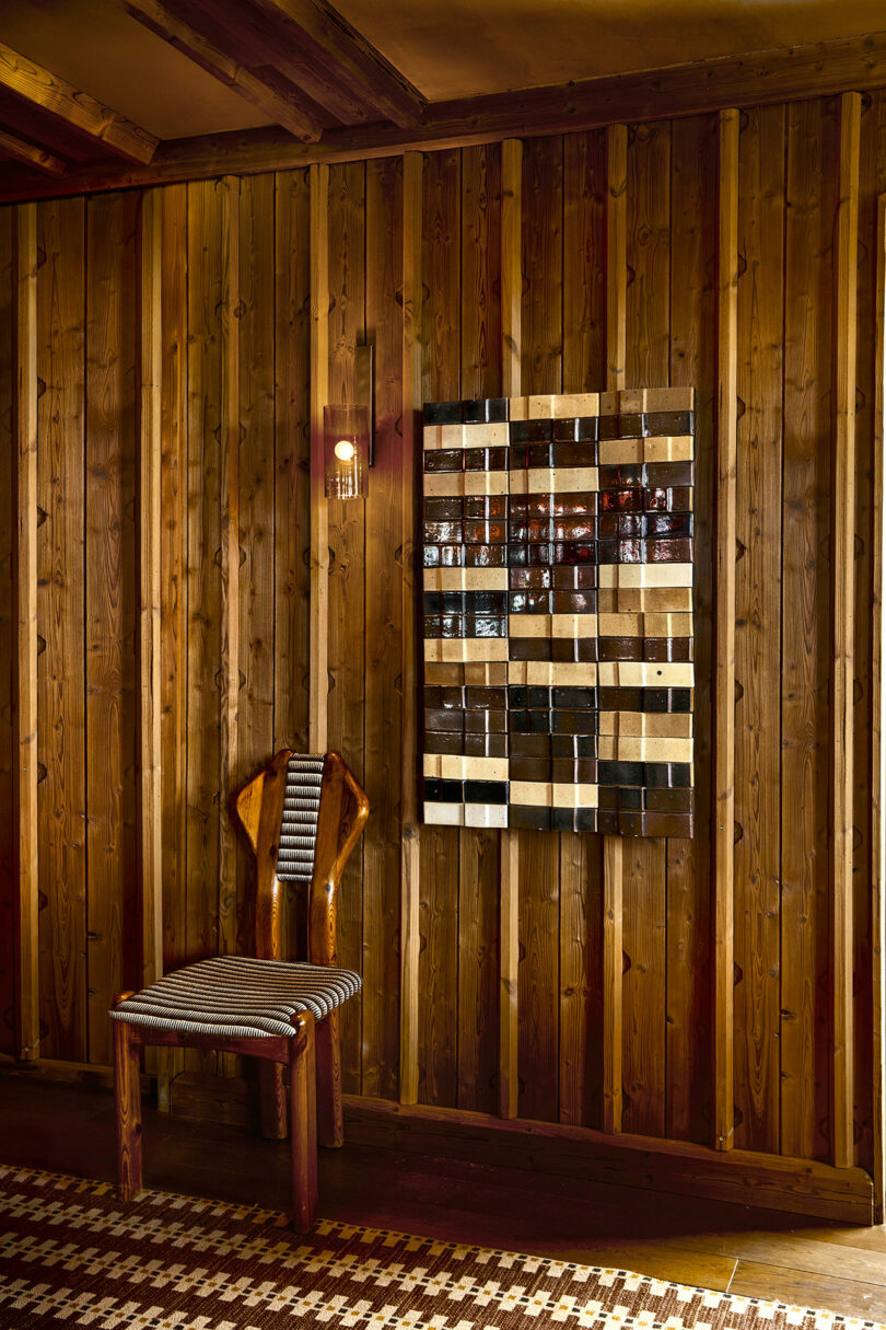 A wooden chair with a striped seat sits against a wood-paneled wall, beside a sconce and a geometric wall art piece with brown, black, and beige tiles.