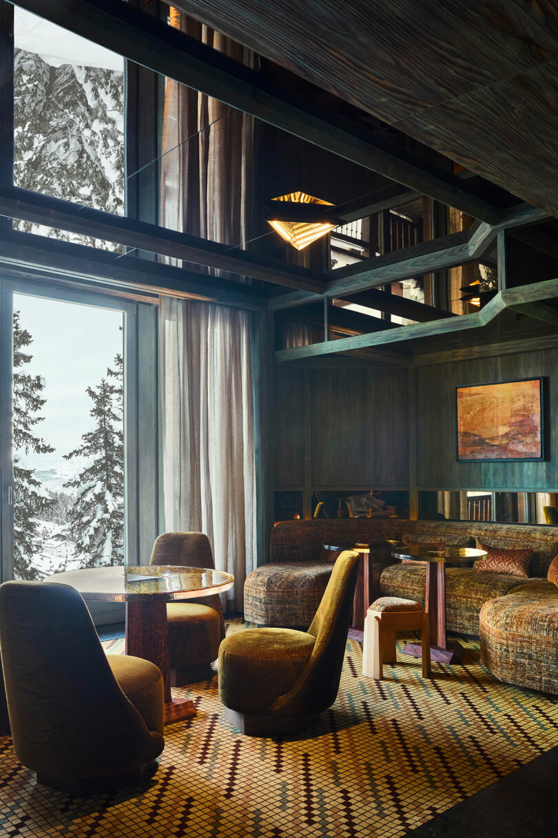 Modern lounge with patterned carpet, round chairs, and a table near large windows overlooking snowy trees and mountains. Warm lighting and wood accents create a cozy atmosphere.