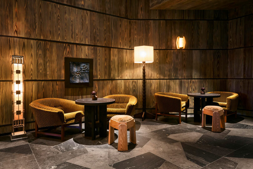 A dimly lit lounge with wooden walls, two seating areas with round tables, cushioned chairs, wooden stools, and three floor lamps on a patterned stone floor.