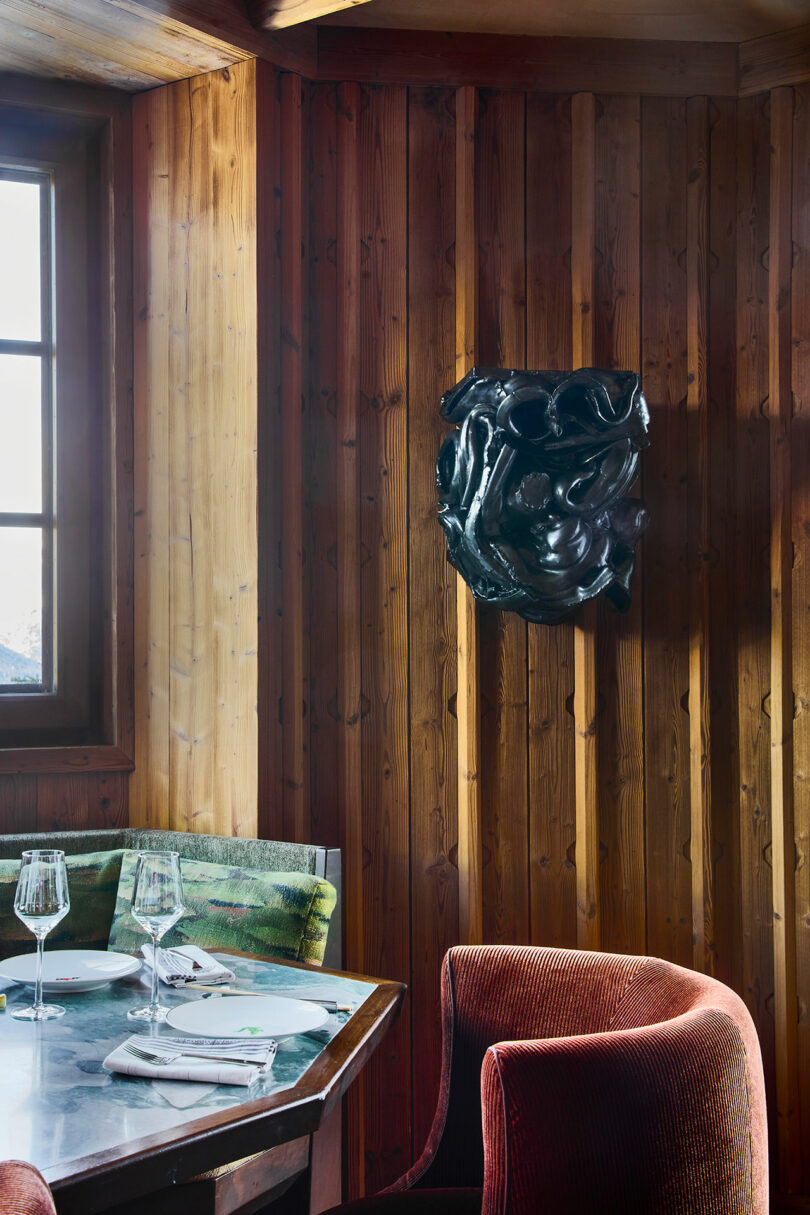 A small dining table set for two stands by a wood-paneled wall with a black abstract sculpture mounted above, next to a window with natural light.