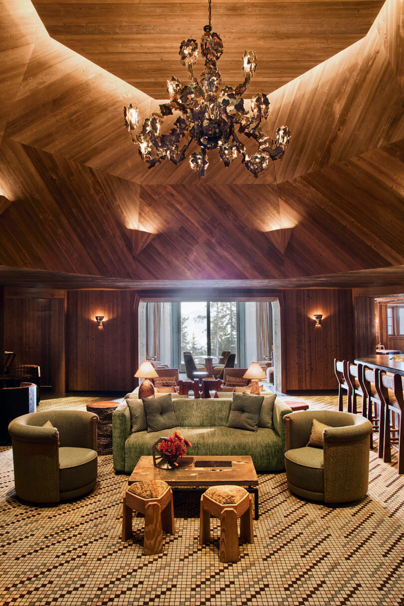 A spacious living room with wood-paneled walls and ceiling, a large chandelier, green sofa, round chairs, tiled floor, and a view of trees through sliding glass doors.