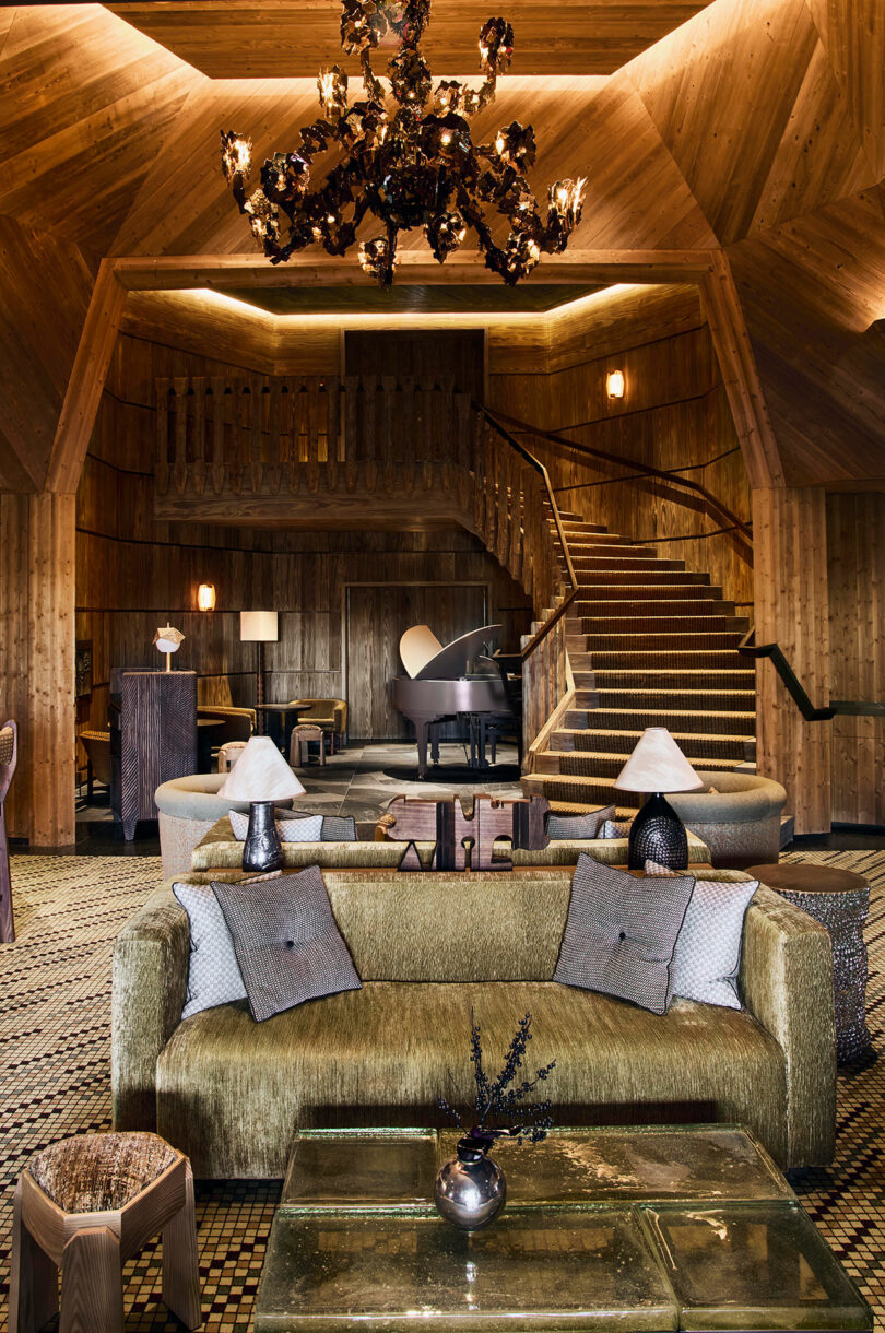 A wood-paneled lounge with a gold sofa, glass coffee table, piano, and a black chandelier, featuring a staircase and upper loft area in the background.