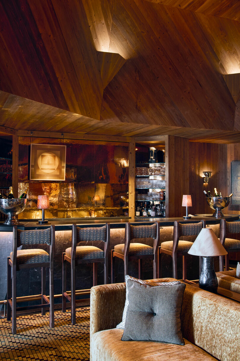 A cozy bar area with wooden walls and ceiling, a row of barstools at a counter, small lamps, and shelves with glasses and bottles behind the bar. A sofa with cushions is in the foreground.
