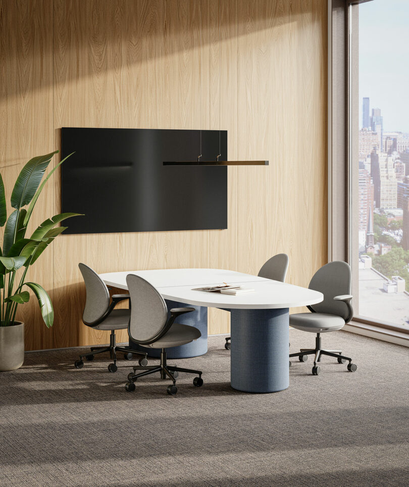A modern KFi STUDiOS conference room with a white oval table, four gray chairs on casters, a large black wall panel, a potted plant, and a floor-to-ceiling window overlooking a cityscape.