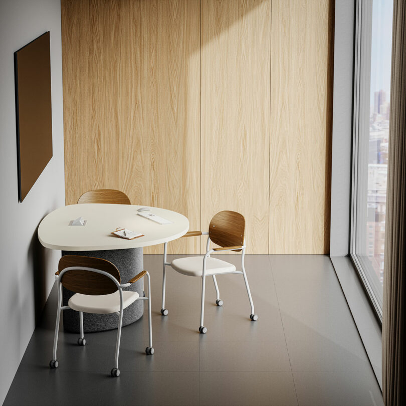 A small KFi STUDiOS meeting area features a white oval table, three wooden chairs on wheels, a wall-mounted board, and a large window overlooking the city.