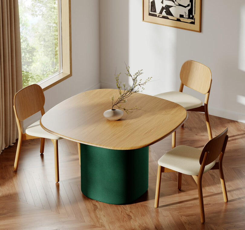 A modern dining area by KFi STUDiOS features a wooden table with a green base, three wooden chairs with light cushions, a vase with branches, and sunlight streaming through a large window.