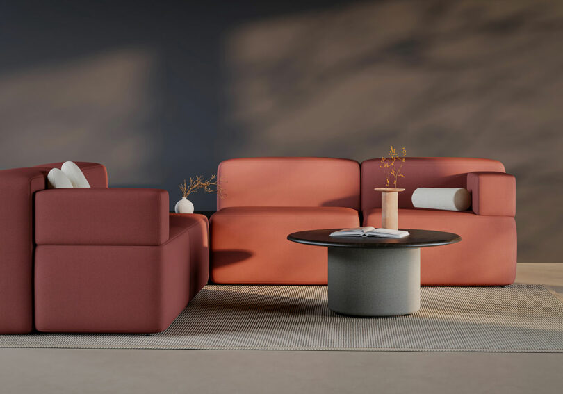A modern living room by KFi STUDiOS features modular red sofas, a round coffee table with a vase and book, and a neutral rug set against a dark accent wall.