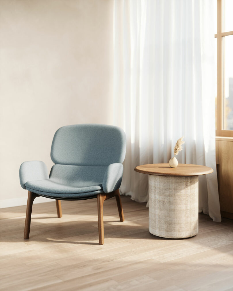 A modern KFi STUDiOS lounge chair with wooden legs sits next to a round side table with a small vase, in a sunlit room with light curtains and wooden flooring.