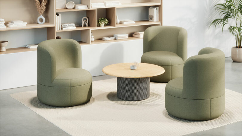 Three green upholstered chairs by KFi STUDiOS surround a round wooden coffee table on a beige rug in a modern, minimalist room with shelves and decorative items.