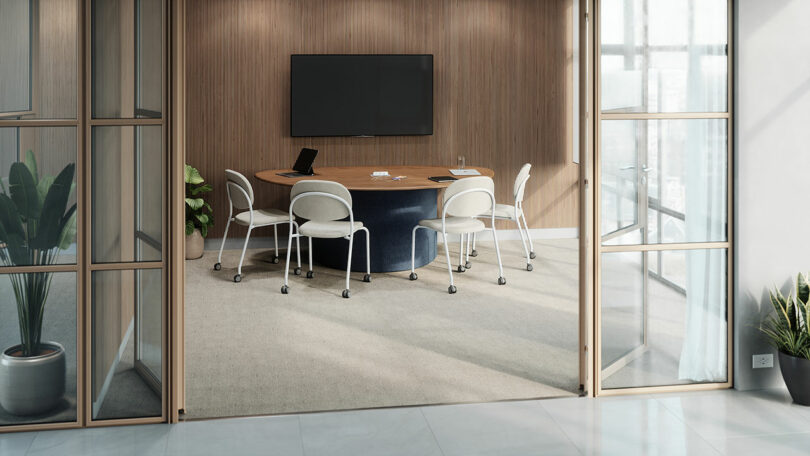 A modern conference room by KFi STUDiOS features a round table, six white chairs, a wall-mounted screen, a laptop, and natural light streaming through large windows.