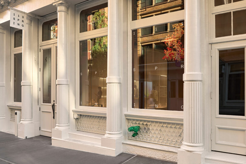 Street-level view of a white building facade with large windows, decorative columns, and a sign reading "113 SPRING." A green hydrant cap is visible near the base.