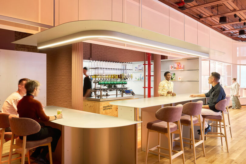 Four people sit and stand around a modern office bar area with stools, wood flooring, and a shelf of bottles in the background.